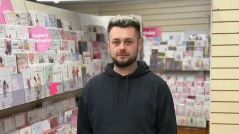 Callum McCall has short black and white hair. He is wearing a black hoody and is standing inside a card shop with greetings cards on display in the background.