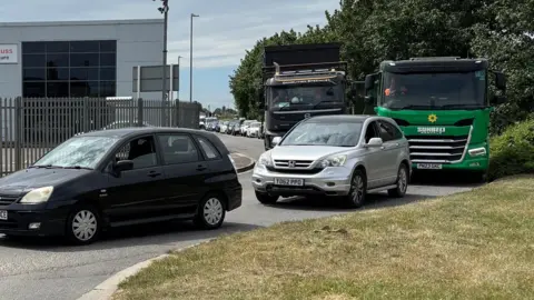 Cars and trucks of various colours queuing in a narrow street on an industrial estate. The queue stretches back as far as we can see. In the foreground is a patch of green grass. In the background are green trees.