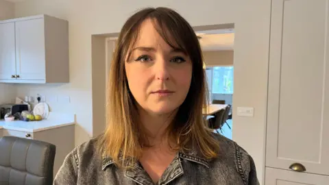 Crystal pictured indoors in her kitchen; she's wearing a grey-black denim jacket and is looking directly into the camera with a sombre expression.
