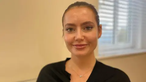 Lydia is wearing a black top and is smiling at the camera. Her dark hair is pulled back into a bun and she is sat in a room with cream walls and a window behind her. It is a head and shoulders shot. She is in focus while the background is slightly blurred.
