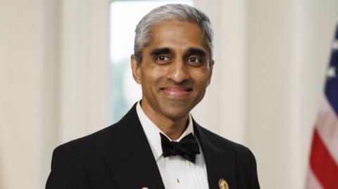 US Surgeon General Vivek Murthy smiling, wearing a sharp suit at a presidential event in the US