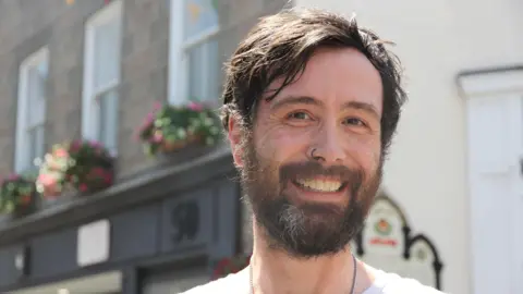 Clinton Millard is a man with dark hair and a dark beard with a grey streak. He smiles at the camera. He was a black nose ring and brown eyes. Behind him is a grey and black high street building with pink and green flower pots. 