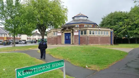 A library building that has a blue metal gate and several small windows. In front of it is grass as well as concrete paths and two large trees. A police officer in black uniform is walking towards it