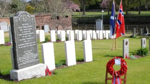 SNC A cemetery with Scotland and Norway flags intertwined and gravestones across the grass and a wreath of poppies with Norwegian colours attached