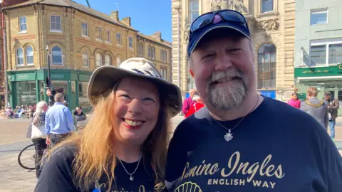Sonja Davison is wearing a hat and has shoulder length ginger hair. Jon is stood on the right, wearing a cap with sunglasses resting on it. Both are wearing blue T-shirts that say Camino Ingles/English Way.