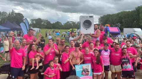 SSNAP In this group photo the runners are joined by partners and children. They are clapping and cheering. The washing machine is held aloft triumphantly.