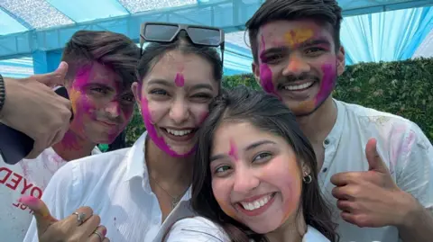 TheBiGhitentertainment Two women and two men wearing white tops are covered with bright pink powder as part of the Holi celebrations. They are all smiling.
