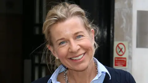Katie Hopkins smiles at the camera with her head tilted to her right shoulder. She is wearing a navy blue jumper with a blue shirt underneath.