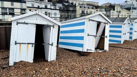 A line of beach huts all scattered across a stony beach and not where they are supposed to be. They are blue and white. The pier can be seen in the back ground. 