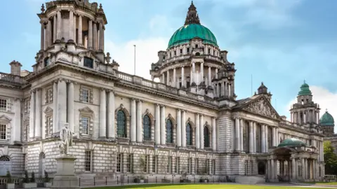 Getty A grand looking white washed building with pillars, beautiful blue stained window glasses and deep panes and a teal blue roof with spires.
Outside on the grounds grass is visible and there is a statue of a man standing on the right.