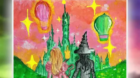 BBFC The artwork itself, which shows the backs of Elphaba, who has long blonde hair and wears a pink ballgown, and Glinda, with her pointy black hat and black dress, holding hands. They look on to the Emerald City, which has green towers and structures and hills sloping down on both sides. Two hot air balloons - one pink and the other green - glide above through a pinky-orange sunset with yellow stars shining in the sky.