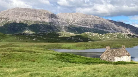 A small, stone cottage sits beside a tranquil loch in an open, grassy landscape. Behind it, rugged grey mountains rise under a sky filled with mixed sunshine and clouds, creating a striking Highland-style rural scene.