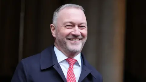 PA Media Government minister Steve Reed, wearing a dark coat and red tie, smiles as he has his photo taken in the street