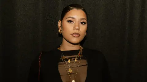 Renait Reborn Milena Sanchez, pictured with her hair tied back in a slick bun. She looks at the camera with a serious expression, wearing a black sheer top and multiple gold necklaces. She's pictured inside against a dark backdrop. 