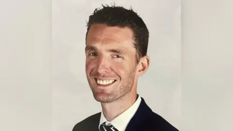 Andrew Burns A head shot of Andy burns. He is smiling in front of a beige background, wearing a black suit jacket, white shirt and a black and white striped tie.