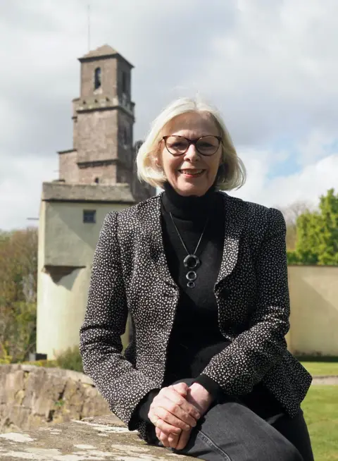 Norman Mackenzie A woman with grey blonde hair and glasses in a dark speckled jacket with a dark top and dark jeans on sits on a wall in front of a Scottish castle.