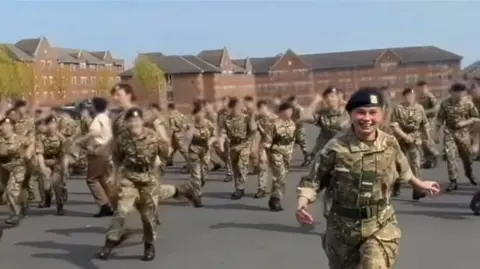 Family Handout A screenshot from a video showing a regiment practicing their uniform marching and saluting on camp at Larkhill. There is a large group of around 30 people dressed in green and brown camouflage uniforms and black caps. They are standing on a large tarmac area surrounded by fields and trees. In the background there is a brown brick building with lots of windows.