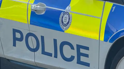 BBC A close-up of the outside of a Guernsey Police car. The car is wrapped in police yellow and blue branding. It has the words POLICE and Guernsey Police logo prominent on the door. 