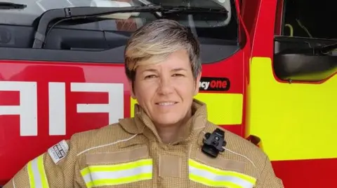 Sally Hammond stands in her firefighter uniform in front of a fire engine. She has short blonde hair and smiles at the camera. 