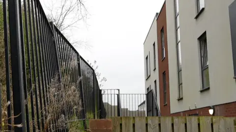 A gate with overgrown bushes leading to a block of flats - where Liam owns a two-bed flat