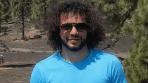 Drew McCormick with long curly brown hair and beard wearing sunglasses and a cyan coloured t-shirt. He is standing in front of trees and hills. He is smiling.