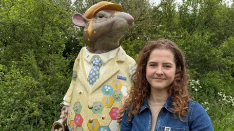 Jess Perrin is pictured standing in front of her Ratty the Water Vole sculpture. She has long, curly dark brown hair and is wearing a denim jumpsuit. She is smiling. Ratty towers over her. He is wearing a yellow flat cap which has honey dripping off it and a colourful suit which has a floral honeycomb pattern. He is holding a briefcase.
