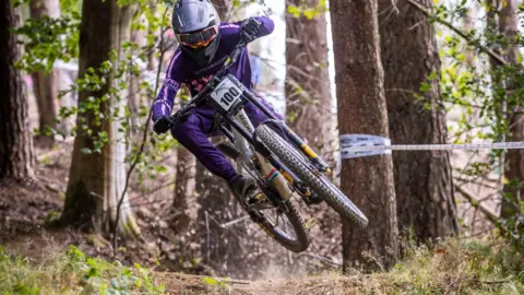A person is mid-air as they ride their bike through the forest. They are wearing a purple outfit, plus a grey helmet and dark sunglasses. There are tall trees in the background. One on the right has white tape wrapped around it.