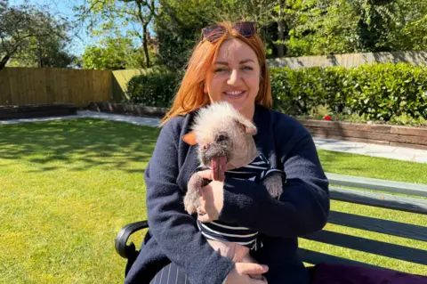 Becki Bowden / BBC A woman with ginger hair holds a striking-looking small dog in her arms while sitting on a green wooden bench in a large, sunny, green garden. The woman is smiling and wears sunglasses on the top of her head, as well as a blue fleece and blue pin-striped trousers. The dog - a pug and Chinese crested cross - has wild, white fur, a long red tongue and big brown eyes. It is wearing a blue and white striped jumper.