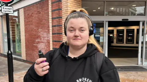 A 26-year-old woman stands in a pedestrian street in front of a high street bank. She is wearing a black hoodie and black headphones and holds a brightly coloured vape in her hand.