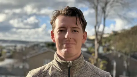 BBC Max Andrews smiles at the camera. He is wearing a grey funnel neck fleece. He has slicked back black hair. St Helier is visible in the background, along with the coastline.