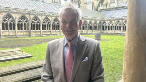 James Turner Lincolnshire County Council leader Martin Hill. He is standing in the grounds of Lincoln Cathedral. He wears a light coloured suit with a light-coloured handkerchief in the breast pocket, a grey shirt and a pink tie. 