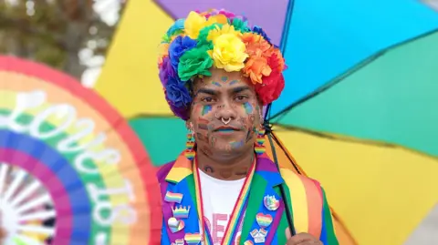 A person with colourful face paint is holding a rainbow umbrella and wearing a rainbow suit. 
