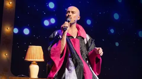 Mark Senior Rob Madge performing in the West End. Madge is wearing a black, pink and crimson outfit while holding a microphone and is sitting on a table with a lamp behind him