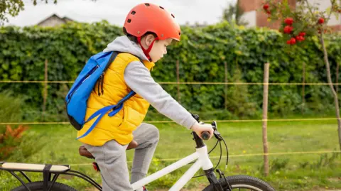 A boy riding a bicycle. He is wearing a yellow gilet, a blue backpack and a red helmet. He is cycling past a green area and is looking to the floor.