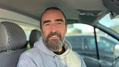 Harry McAlinden is a man with short, black hair and a brown and gray beard. He's wearing a gray hoodie with paint on it. He sits in the cabin of a minibus and smiles at the camera.