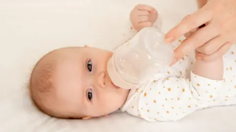Baby wearing a white and yellow babygro lies on its side with a bottle of infant formula to its lips, held there by an adult's hand
