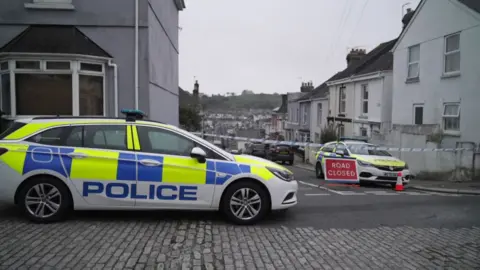 A residential area in Plymouth that has been cordoned off by police. Two police vehicles are parked at the scene, which features one partially cobbled road. A red "ROAD CLOSED" sign and a traffic cone and police tape can be seen stretched across the width of a street to restrict access. The area is lined with terraced houses. 