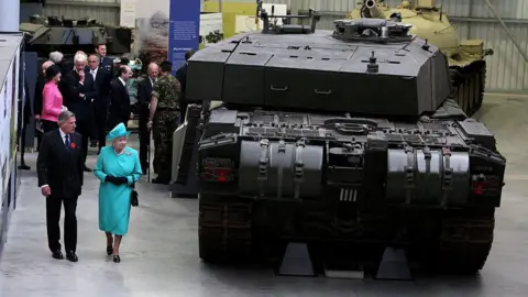 Getty Images The then director, Sir Roger Wheeler shows Queen Elizabeth II around the museum. He wears a dark shut and a red poppy. Her Majesty is dressed in a blue dress and hat. They are walking next to a large tank. 