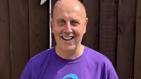 A man stands against a dark fence. He is wearing a purple T shirt and smiling. He has a bald head and grey moustache