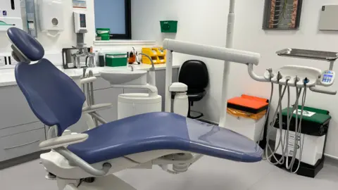 There is a blue dentist chair in the foreground. Other dental equipment can be seen in the background and there are orange and a green waste bins.