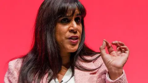Ian Forsyth/Getty Images Close-up of a lady with brunette hair, wearing a white shirt with a pink blazer over the top, gesturing with her hand as she speaks