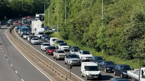 A queue of traffic along both lanes of a dual carriageway heading from the top left to the bottom right of the picture. The traffic includes lorries, cars and vans. There is a low barrier in the centre of the road and some vehicles are visible heading in the opposite direction in the top right. There are trees to the right of the photo and lamp poles.