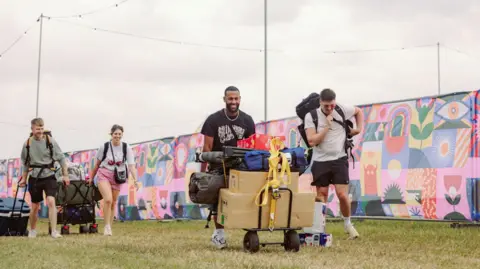 Josh Collins/Truck Festival Four festival goers arrive at the venue, there are all wheeling trunks with their camping gear and luggage, smiling. There is a colourful backdrop sign lining the festival grounds in the background.