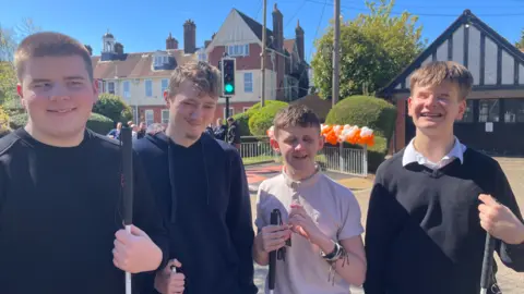 Four people, including three of them wearing dark clothing, are prominent in the photo. Two buildings and railings are behind them.