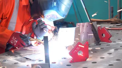 BBC Welder wearing orange protective suit, with mask and gloves on while welding together two pieces of steel