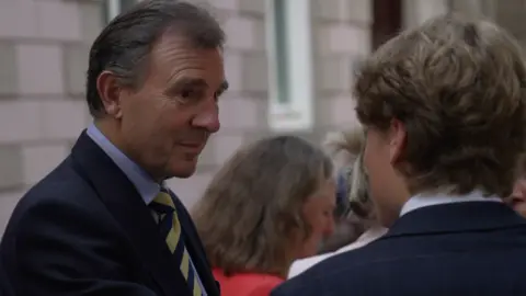 A person in a dark suit and striped tie speaks with another person in a suit at an indoor event, with several people blurred in the background.