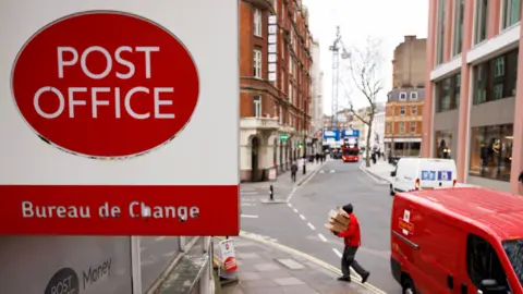 A view of a Post Office sign that reads 'Post Office; Bureau de Change' in red and white with a postal worker delivering boxes from a red Royal Mail van on a street in London in 2024.
