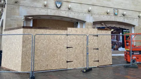 Wooden chipboards covering the entrance to a shop. The boards are behind a metal chain fence. A orange hydraulic lift is to the right.