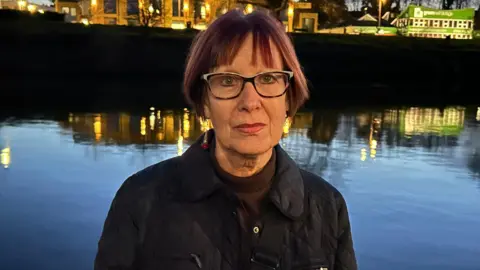 Gavin Kermack/BBC A woman with mid-length straight hair and spectacles stands in front of a river at night, with lit-up buildings in the background.