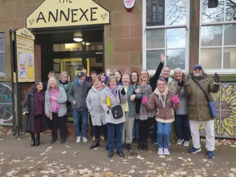 SLF A group of people stand waving and cheering outside a sandstone building with the nameplate The Annexe on it